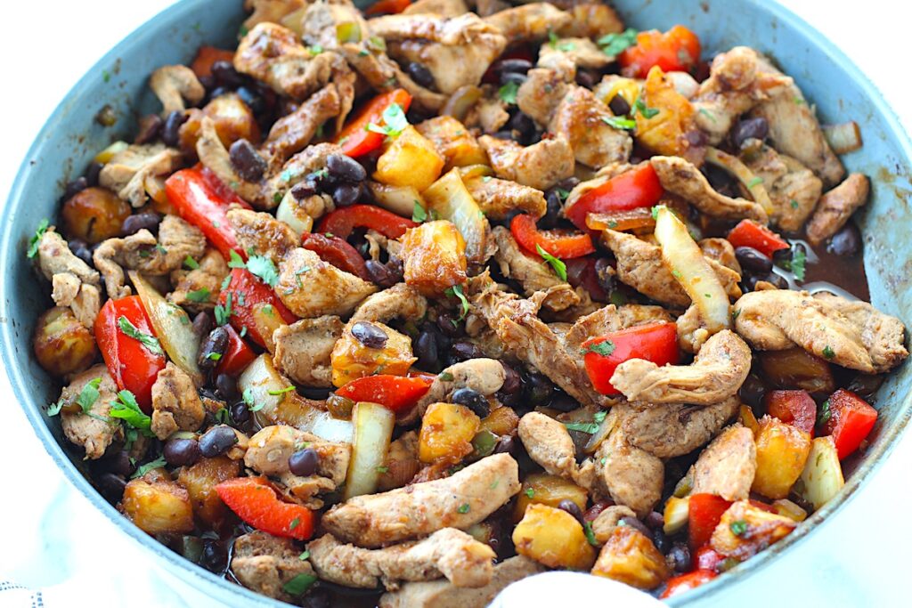 Brazilian Marinated Chicken Stir Fry with pineapple, black beans, red Pepper and onions in a skillet wrapped in a towel with cilantro garnish. It is truly one amazing dish with incredible flavor!