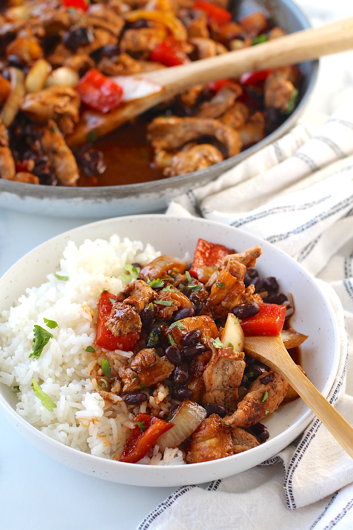 Brazilian Marinated Chicken Stir Fry with pineapple, black beans, red Pepper and onions in a bowl over white rice with a bamboo fork and cilantro garnish.  It is truly one amazing dish with incredible flavor!