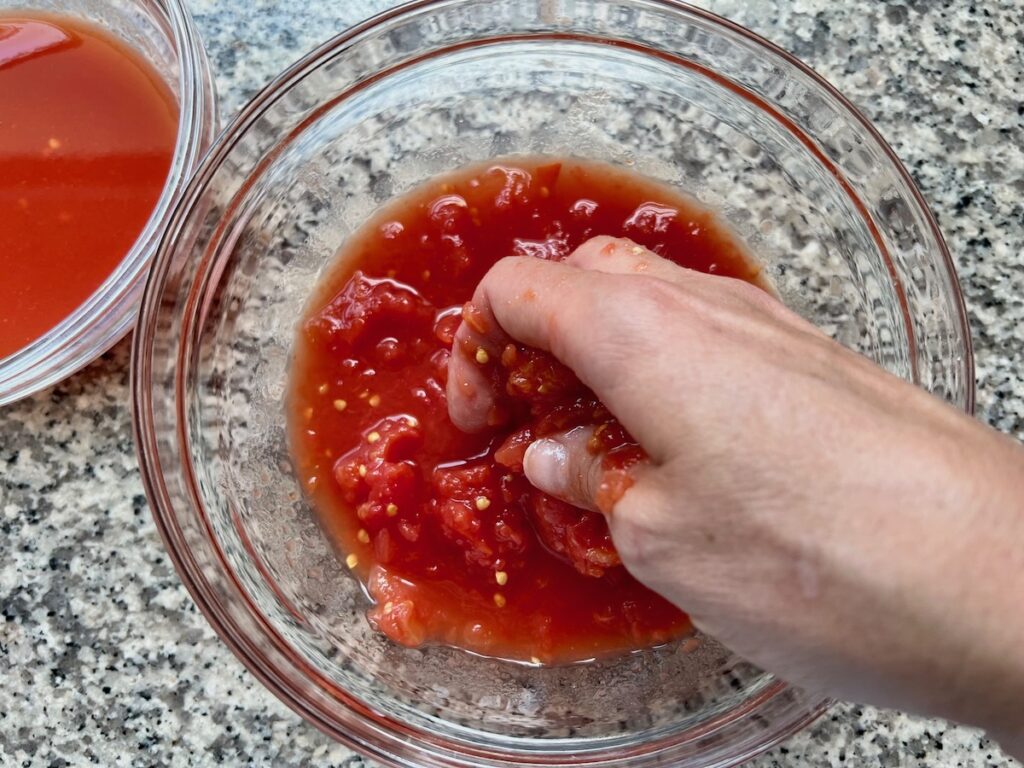 Hand crushing tomatoes in a bowl with tomato sauce in a smaller bowl to the side for Baked Rice with Cheese and Tomatoes.