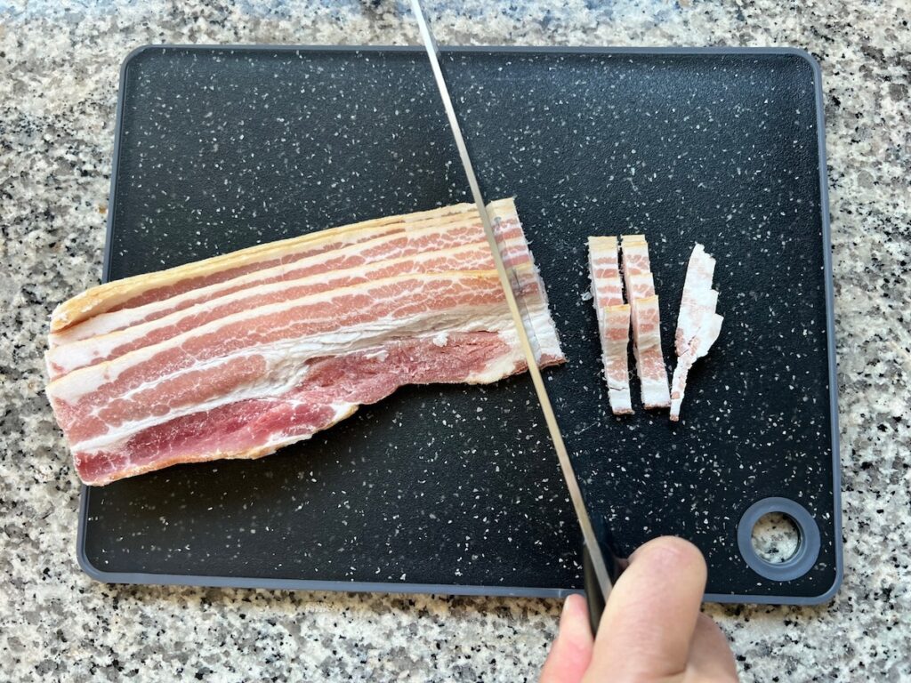 Knife slicing frozen bacon slices on a black cutting board for Carioca Beans recipe.