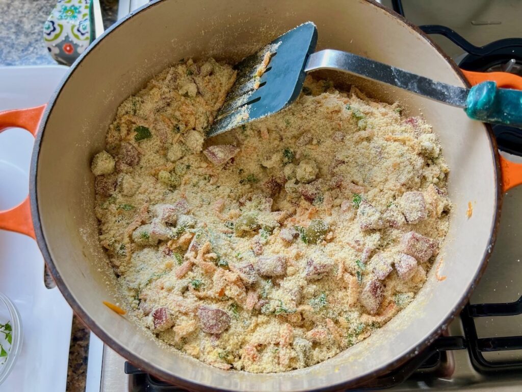 Spatula mixxing in the cassava flour into the sausage, carrots, garlic, bacon, olives and scallions in a dutch oven for Brazilian Farofa Recipe.
