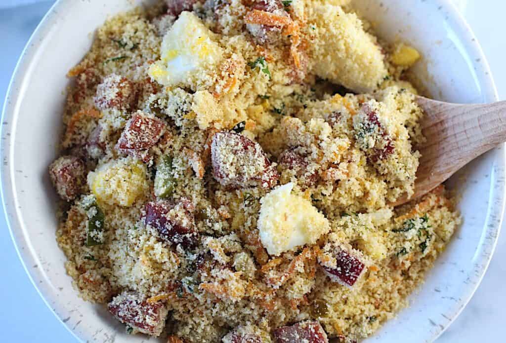 Brazilian Farofa Recipe in a bowl on blue and white towel on counter. It has bacon, sausage, green olives, hard boiled eggs, carrots, onions, and garlic and is a delicious traditional recipe for Christmas ~ Farofa de Natal.