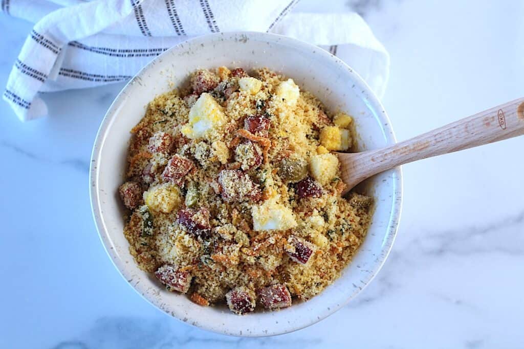 Brazilian Farofa Recipe in a bowl with wood spoon on blue and white towel on counter. It has bacon, sausage, green olives, hard boiled eggs, carrots, onions, and garlic and is a delicious traditional recipe for Christmas ~ Farofa de Natal.