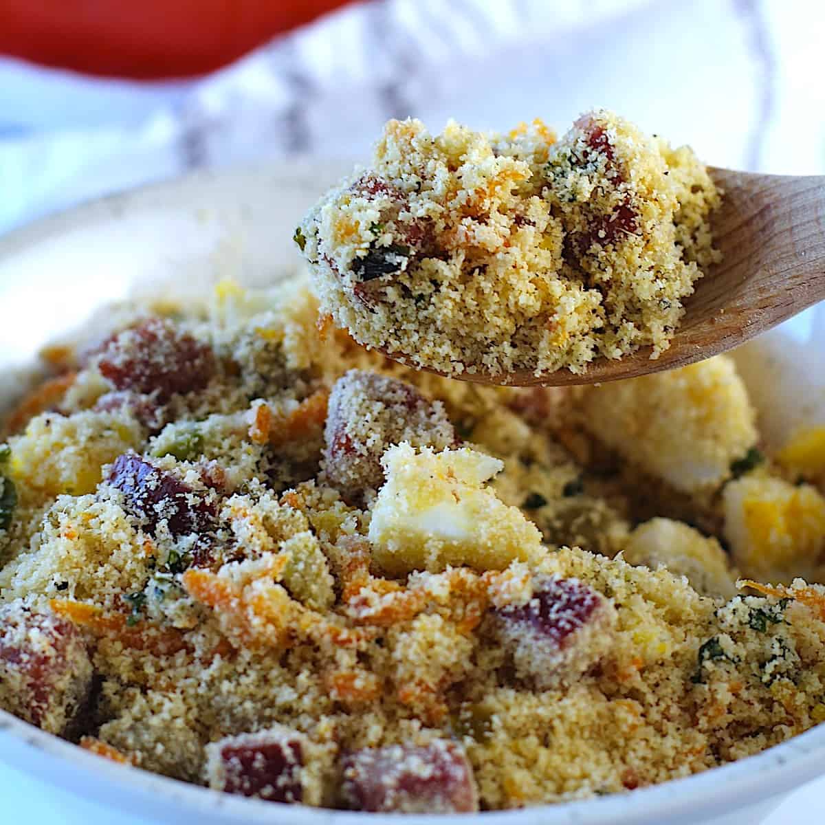 Brazilian Farofa Recipe being scooped up by a wood spoon over a bowl on blue and white towel on counter. It has bacon, sausage, green olives, hard boiled eggs, carrots, onions, and garlic and is a delicious traditional recipe for Christmas ~ Farofa de Natal.