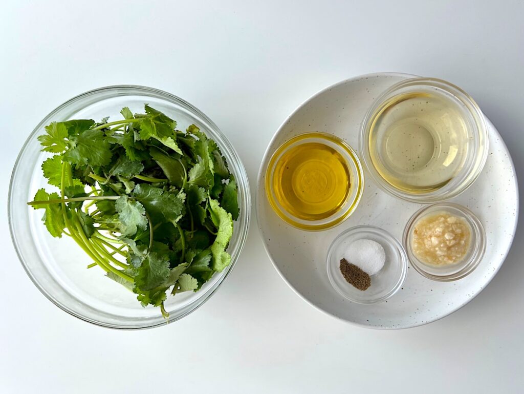 Fresh cilantro leaves in a bowl next to vinegar, olive oil, minced garlic, salt, and pepper all measured out into bowls for Brazilian Cilantro Chimichurri recipe.