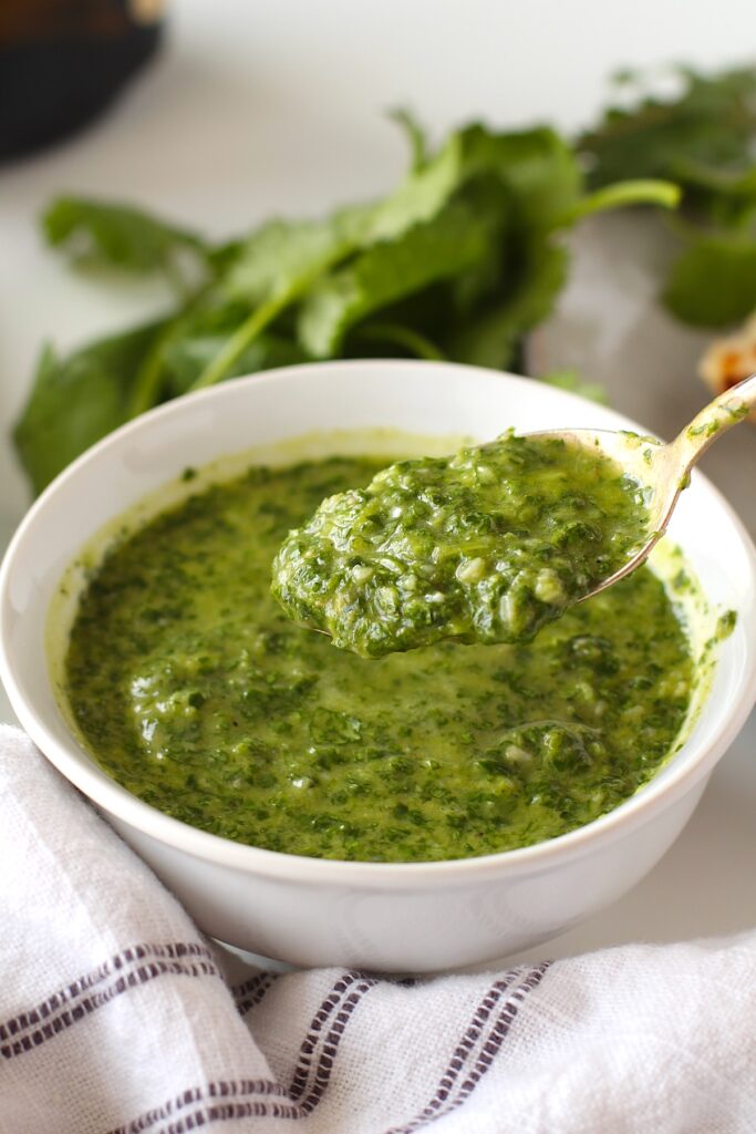 Spoon scooping Brazilian Cilantro Chimichurri recipe over bowl with fresh cilantro and bottle of olive oil in background.
