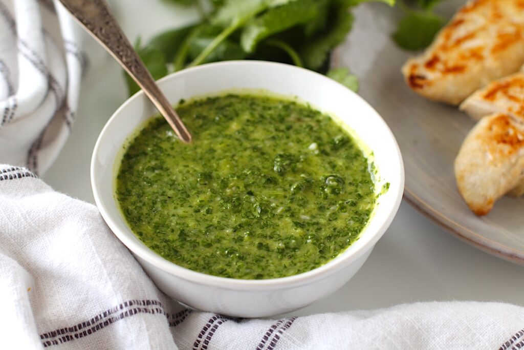 Spoon in bowl of Brazilian Cilantro Chimichurri recipe with fresh cilantro and grilled chicken in background.