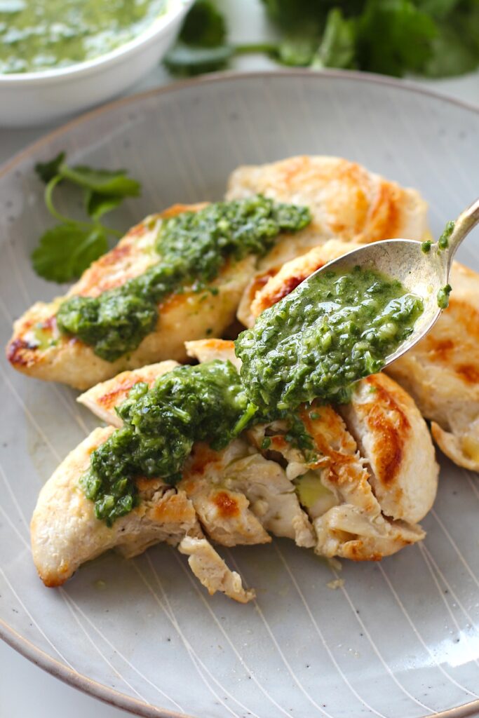 Spoon adding Brazilian Cilantro Chimichurri recipe on top of grilled chicken on a plate.