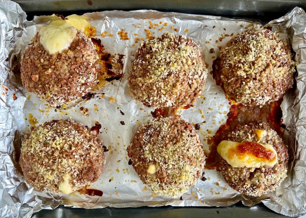 Six cooked breaded polpettone beef patties on an aluminum lined sheet pan with cheese oozing out of two patties.