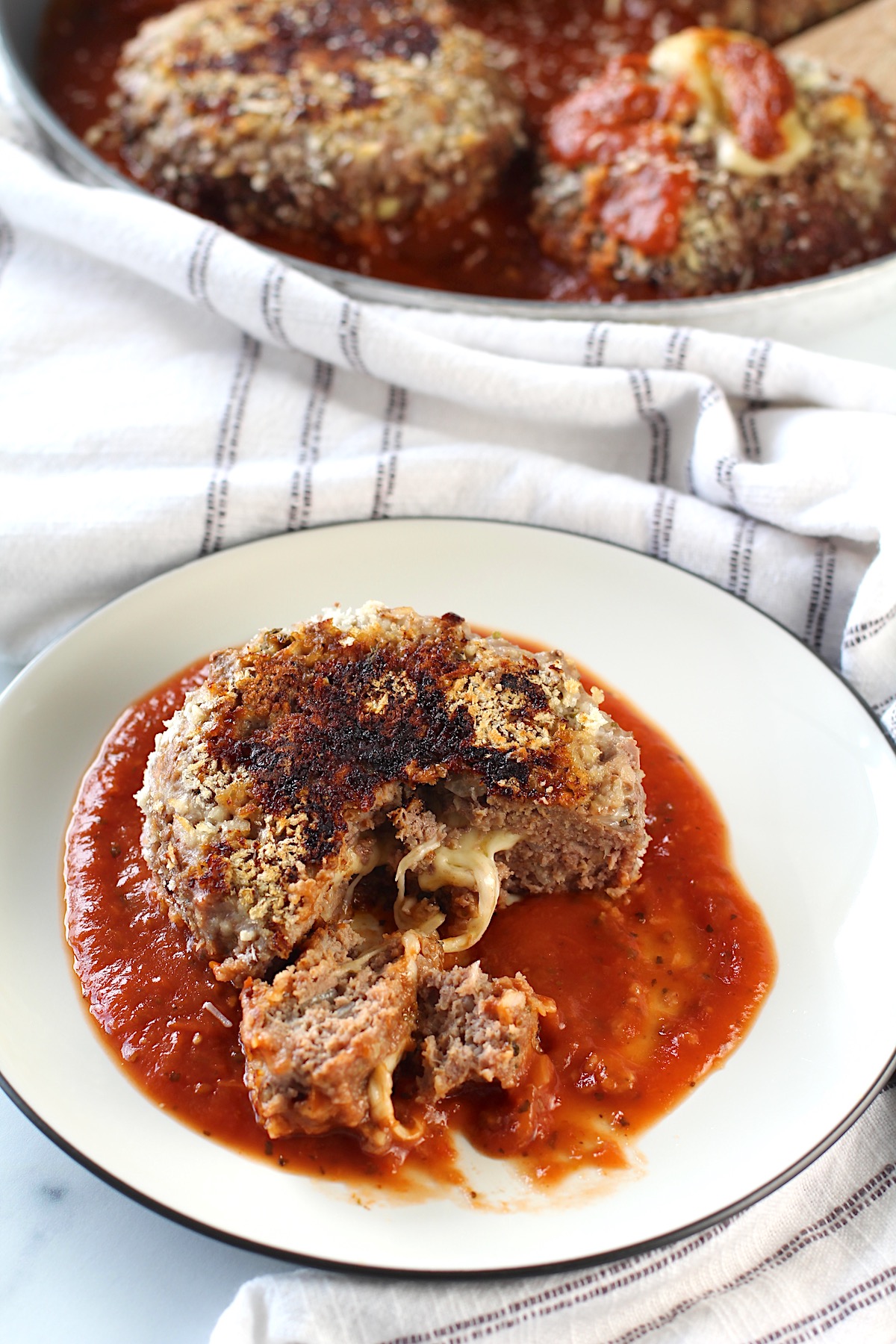A Ground beef Polpettone sitting on tomato sauce on a plate with a piece cut out of it and mozzarella cheese oozing out.  In the background, a skillet with more Polpettone in tomato sauce.