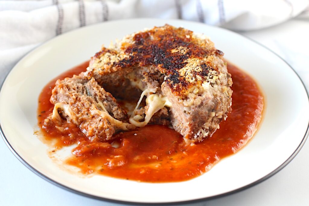 A Ground beef Polpettone sitting on tomato sauce on a plate with a piece cut out of it and mozzarella cheese oozing out.