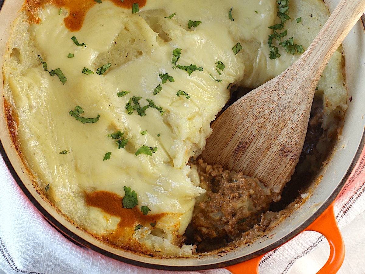 Dutch oven shepherd's pie with ground beef filling topped with mashed potatoes in a dutch oven with wooden spatula resting in place where some pie has been taken out.
