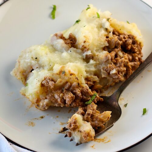 Piece of Dutch oven shepherd's pie with ground beef filling topped with mashed potatoes on a plate with fork laying down with a bite.