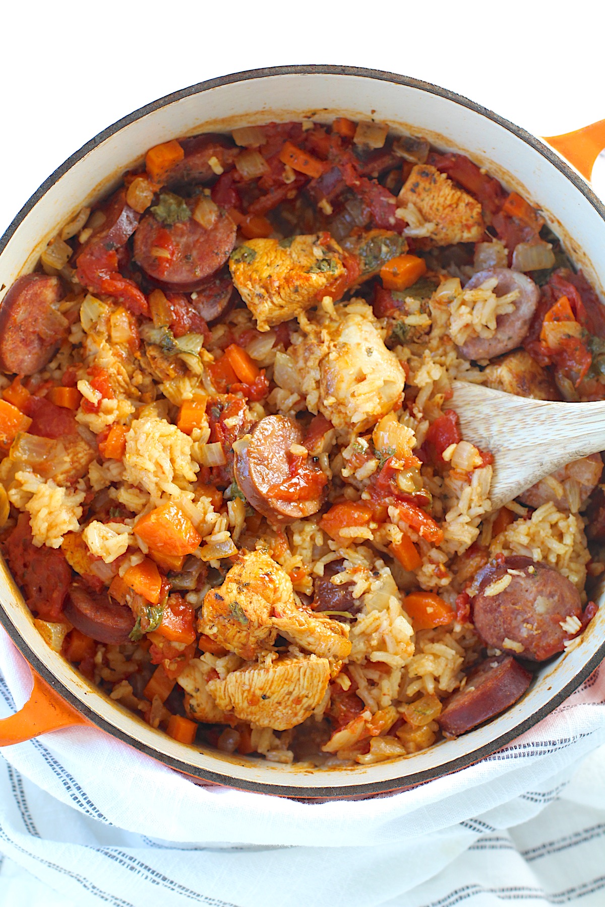 Galinhada, Dutch Oven Chicken and Rice with slices of sausage, carrots, onions, and more in a dutch oven with wood spatula on counter with blue and white towel.