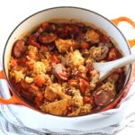 Galinhada, Dutch Oven Chicken and Rice with slices of sausage, carrots, onions, and more in a dutch oven with wood spatula on counter with blue and white towel.