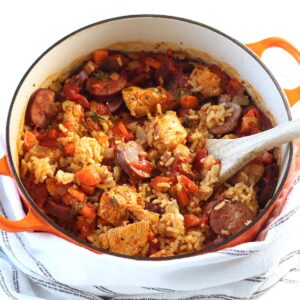 Galinhada, Dutch Oven Chicken and Rice with slices of sausage, carrots, onions, and more in a dutch oven with wood spatula on counter with blue and white towel.