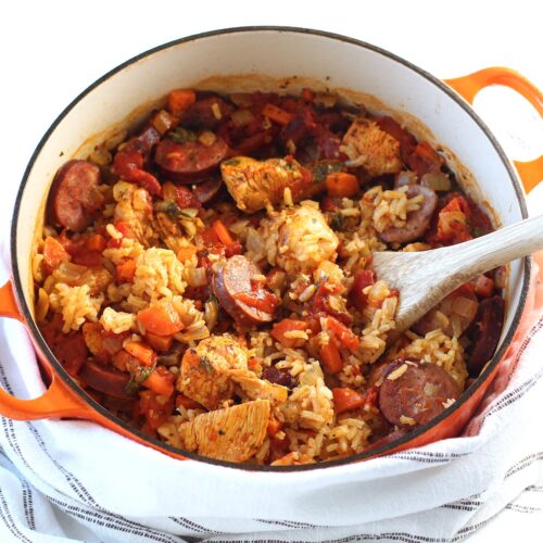 Galinhada, Dutch Oven Chicken and Rice with slices of sausage, carrots, onions, and more in a dutch oven with wood spatula on counter with blue and white towel.