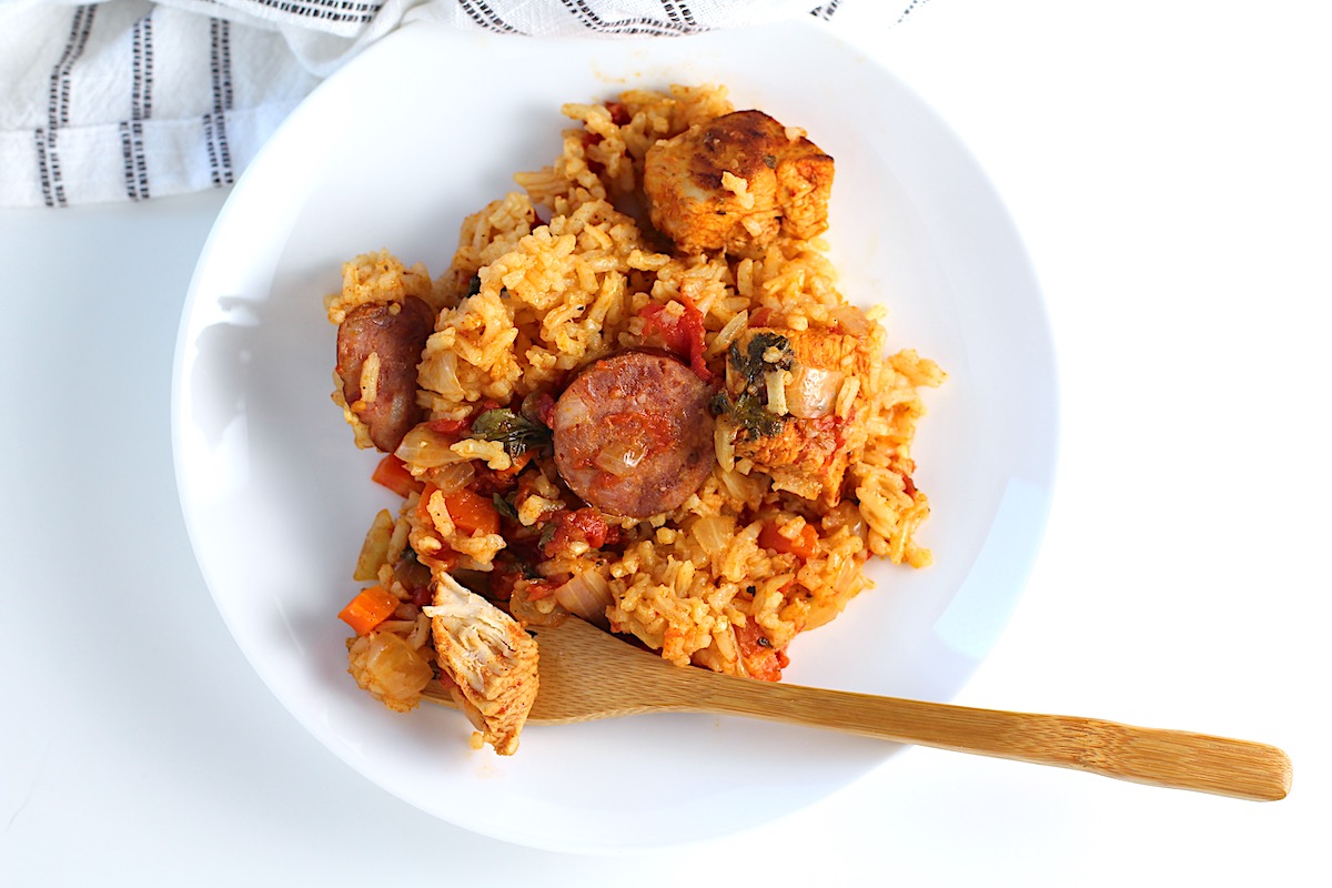 Galinhada, Dutch Oven Chicken and Rice with slices of sausage, carrots, onions, and more on a plate with bamboo work holding a bite of chicken.
