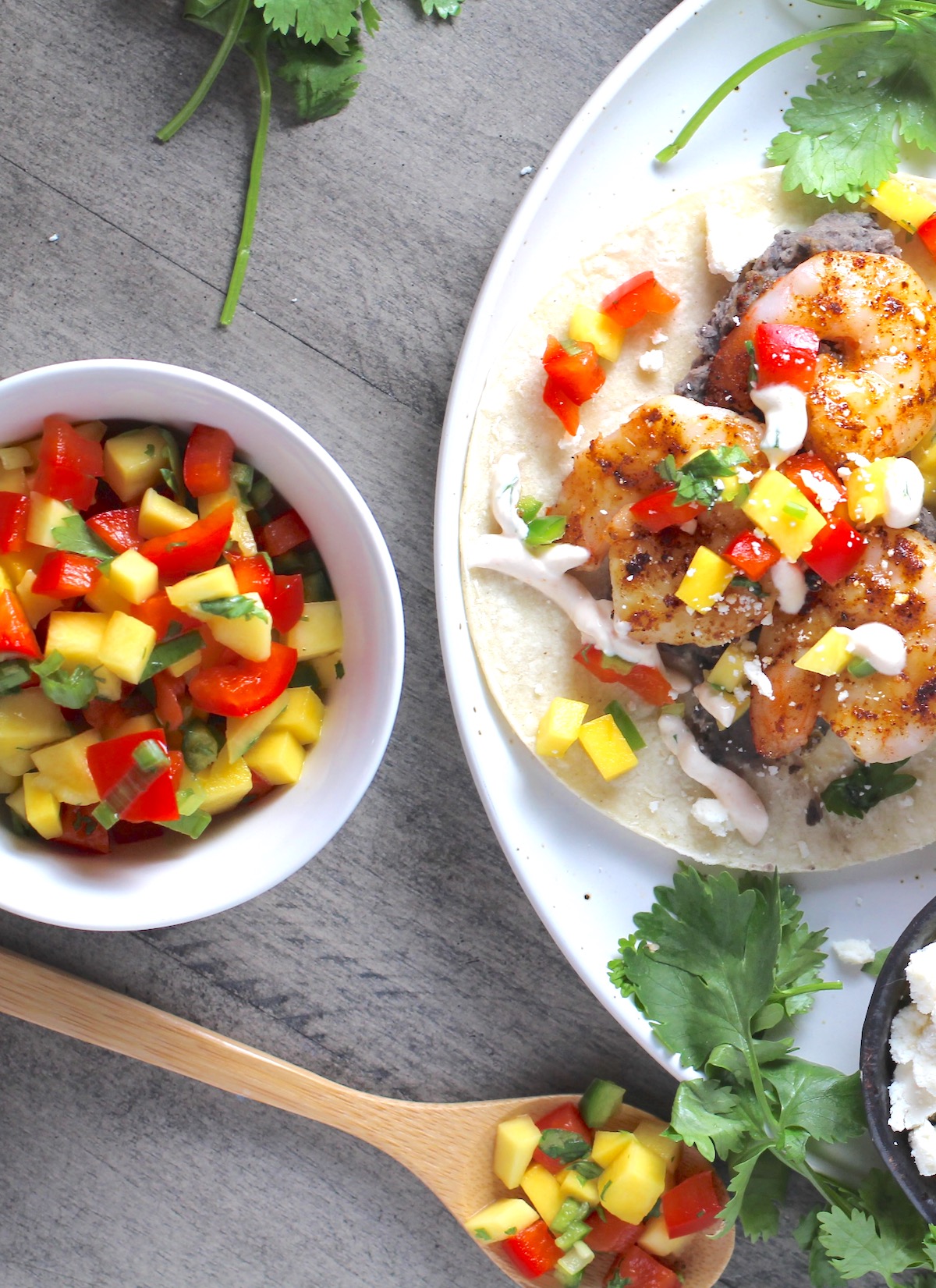 Spicy Mango Salsa in a bowl with a bamboo spoon laying down with a scoop of the salsa and shrimp taco on a plate with the mango salsa on top.