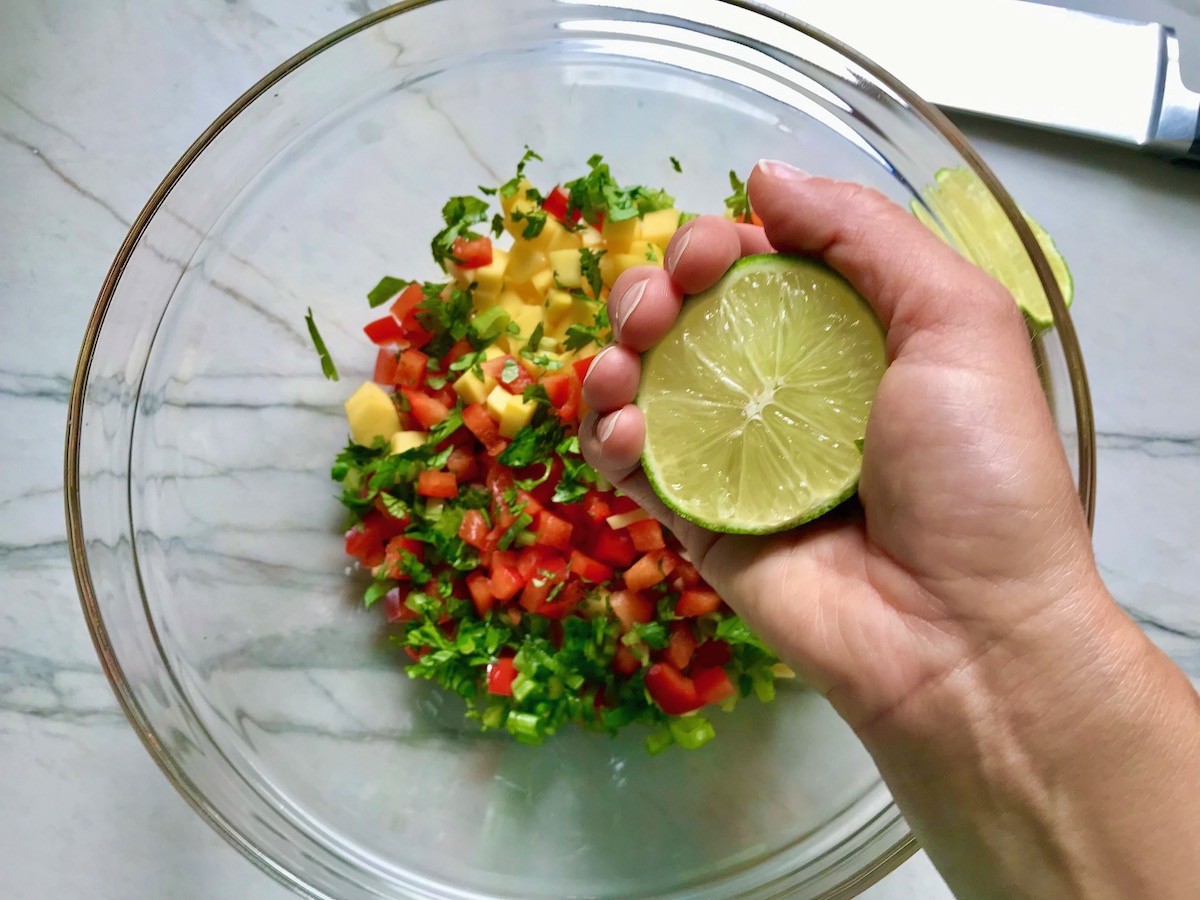 Hand squeezing lime over mango and other ingredients in a bowl for Spicy Mango Salsa.
