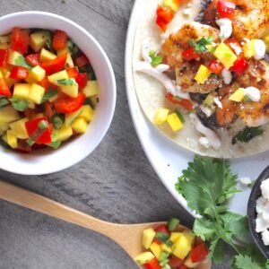 Spicy Mango Salsa in a bowl with a bamboo spoon laying down with a scoop of the salsa and shrimp taco on a plate with the mango salsa on top.