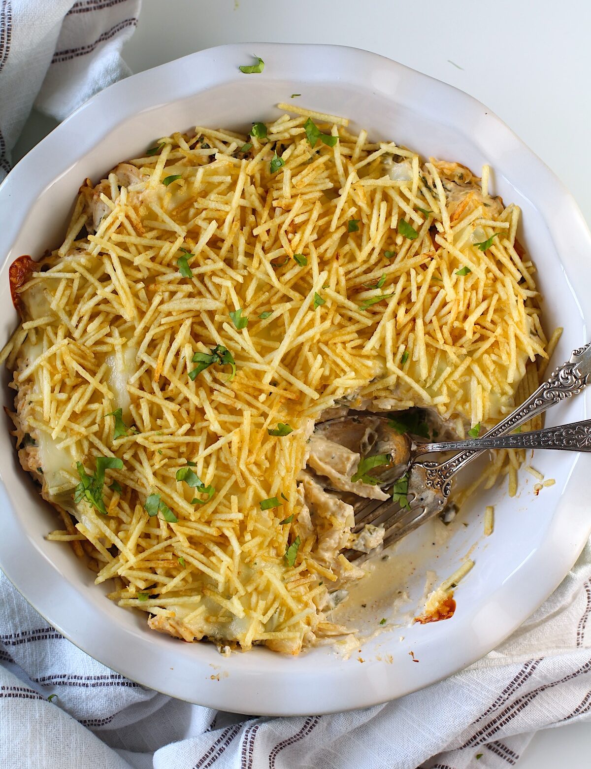 Ultimate Chicken Casserole recipe in a pie dish with a portion removed and a fork and spoon sitting in the dish. It has shredded chicken, green peppers, onions, garlic, cilantro, paprika, and cream cheese holding it all together. On top, you get a layer of ooey gooey cheese and then crunchy potato sticks!