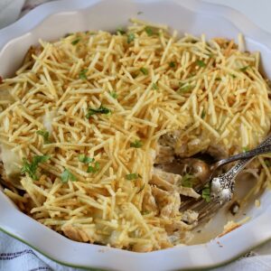 Ultimate Chicken Casserole recipe in a pie dish with a portion removed and a fork and spoon sitting in the dish. It has shredded chicken, green peppers, onions, garlic, cilantro, paprika, and cream cheese holding it all together. On top, you get a layer of ooey gooey cheese and then crunchy potato sticks!