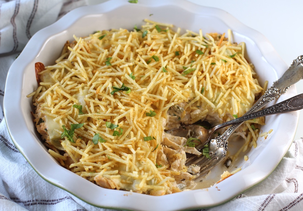 Ultimate Chicken Casserole recipe in a pie dish with a portion removed and a fork and spoon sitting in the dish. It has shredded chicken, green peppers, onions, garlic, cilantro, paprika, and cream cheese holding it all together. On top, you get a layer of ooey gooey cheese and then crunchy potato sticks!