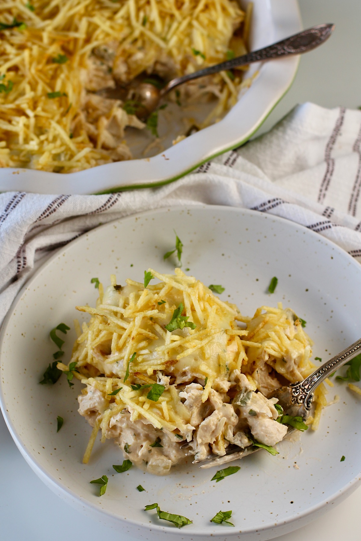 Ultimate Chicken Casserole recipe on a plate with the casserole in the background. It has shredded chicken, green peppers, onions, garlic, cilantro, paprika, and cream cheese holding it all together. On top, you get a layer of ooey gooey cheese and then crunchy potato sticks!