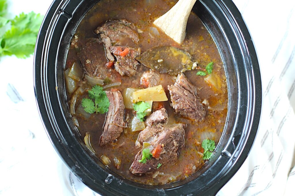 Brazilian Vaca Atolada Beef Rib Stew in slow cooker with wood spoon with yucca, onions, garlic, tomatoes, cilantro in a delicious thick beef wine sauce.