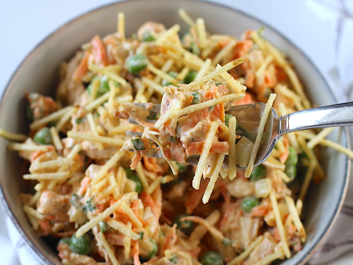 Fork holding up a bite of Brazilian Chicken Salad over the bowl with crispy potato sticks on top.