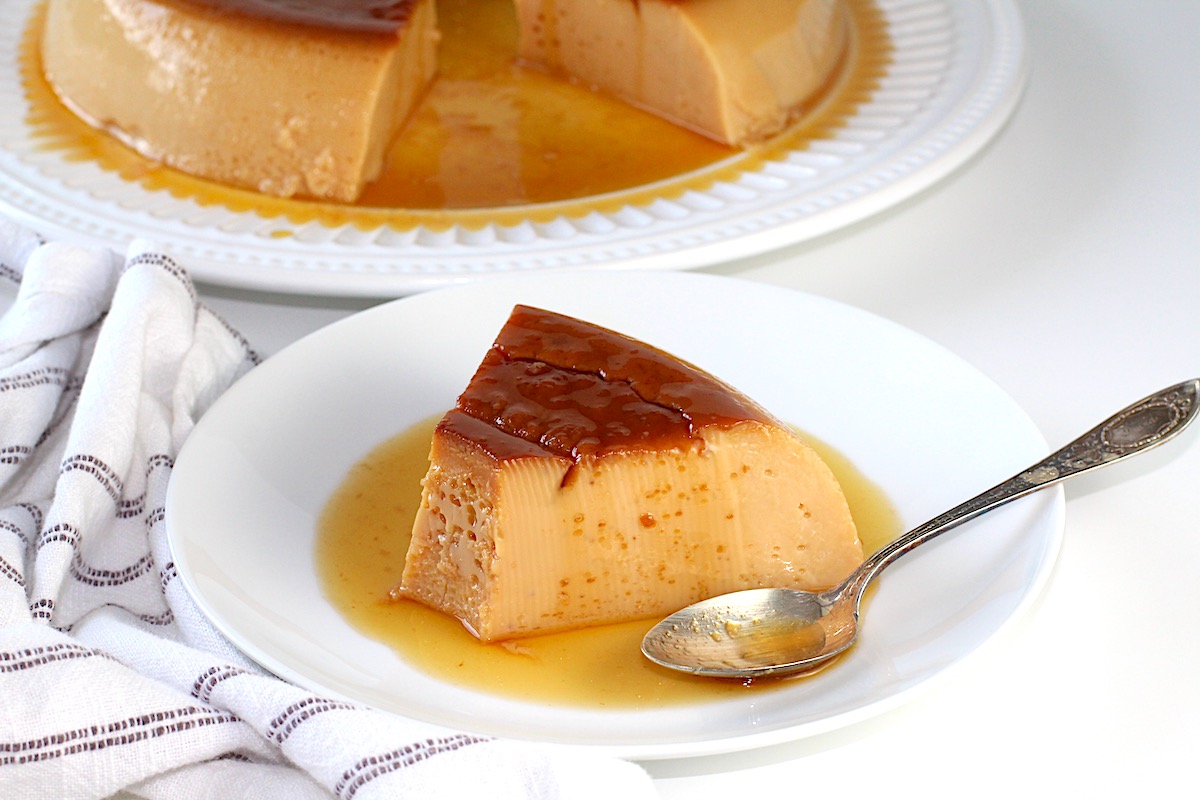 Piece of flan cut and on a plate with a spoon and the rest of the flan on a platter in background for Brazilian Flan Recipe.