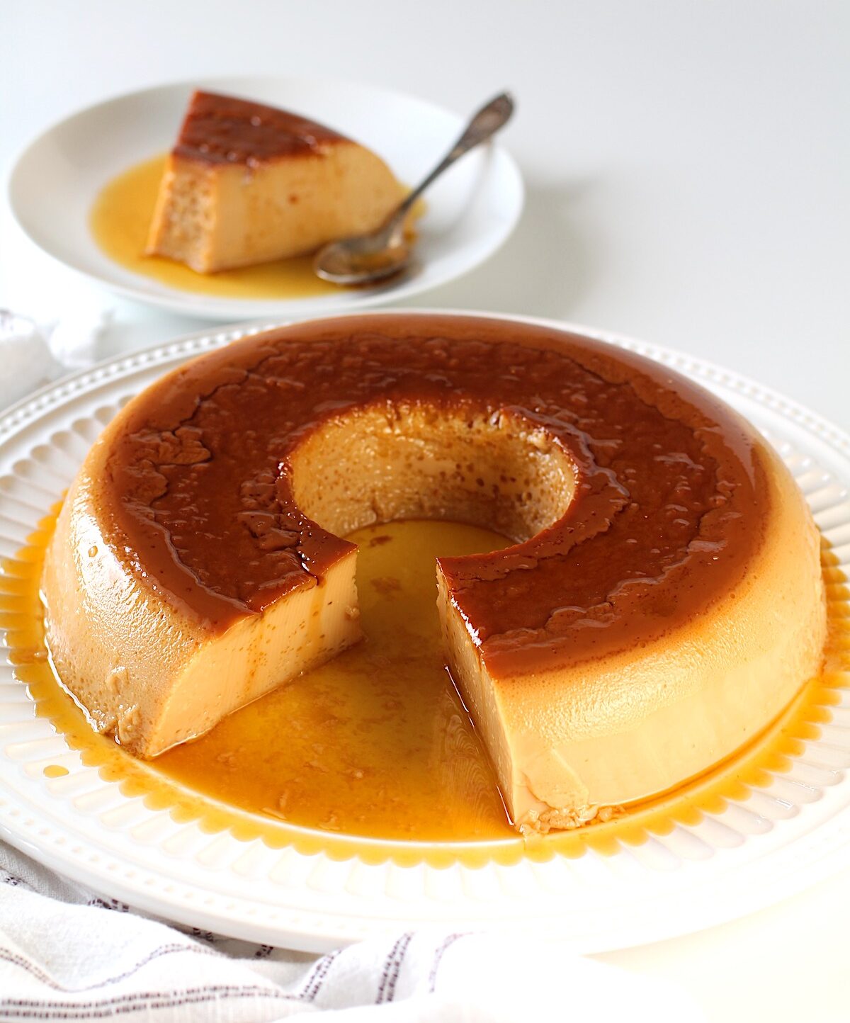 Brazilian Flan recipe in a tube shape with caramel sauce on top on a platter with a piece cut out of the front. In the background is the piece on a plate with a spoon.