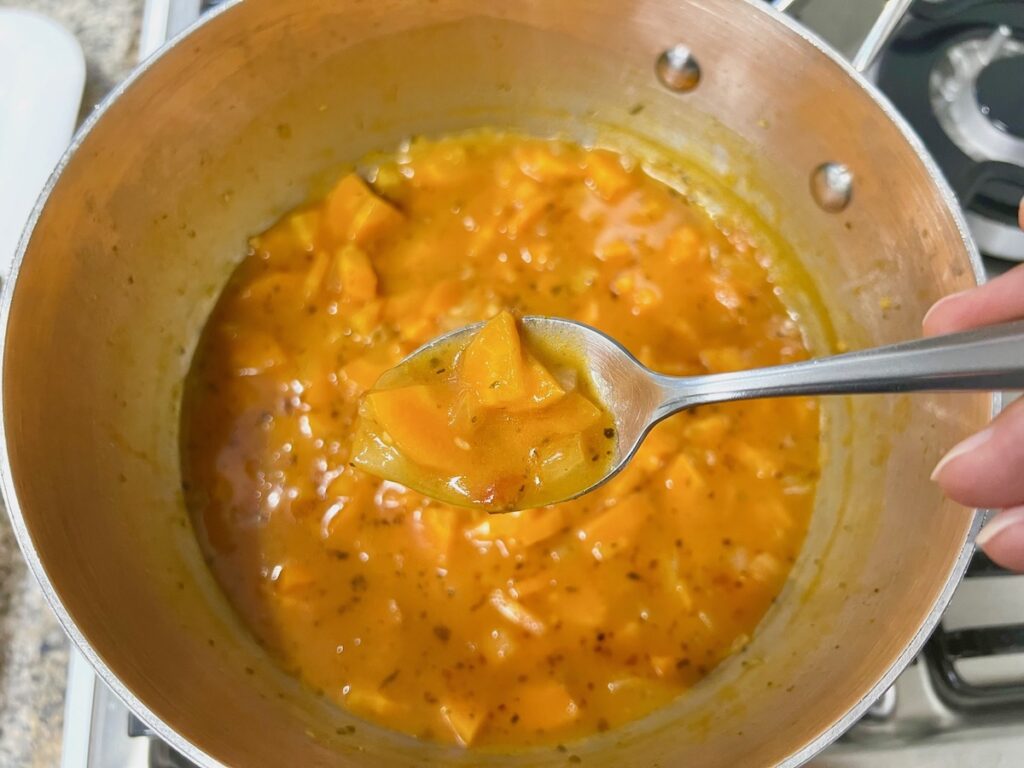 Spoon holding a spoonful of tomato sauce, broth, diced carrots, onions, and minced garlic cooked over a pot for Carrot Sauce.
