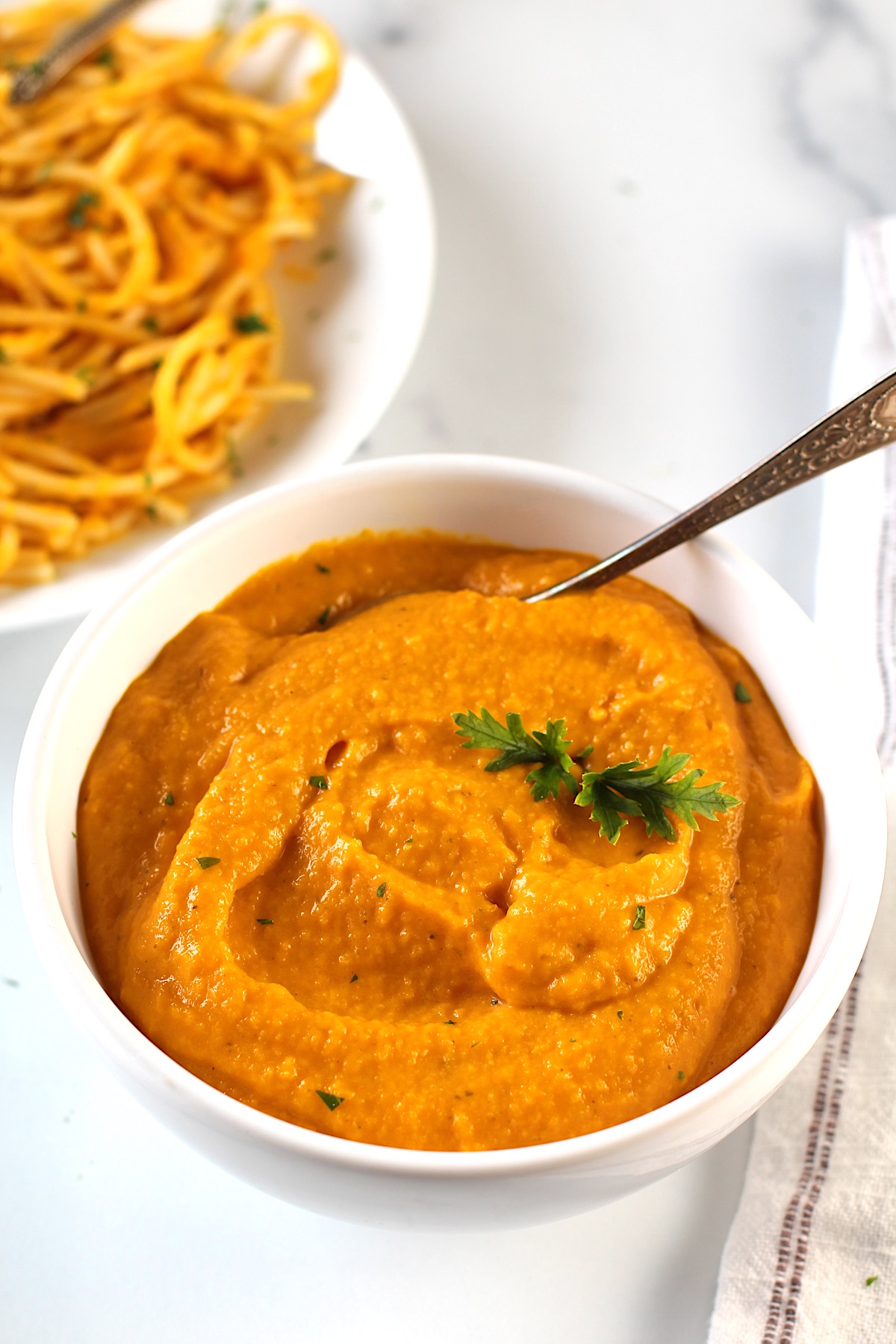 Carrot Sauce in a bowl with a spoon and parsley garnish.  In background is a plate of pasta mixed with carrot sauce and a napkin on counter.