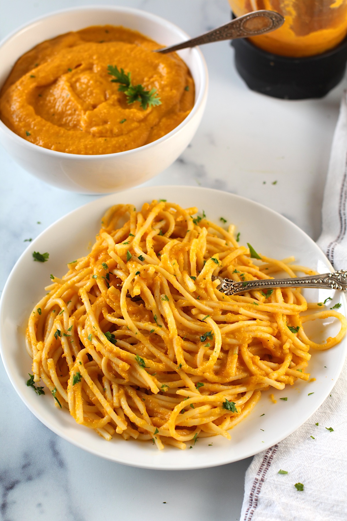 Plate of pasta mixed with carrot sauce with a fork in pasta and a napkin on counter. In the background is a bowl of carrot sauce with a spoon and parsley garnish. Also in background is the blender with the sauce in the bottom.