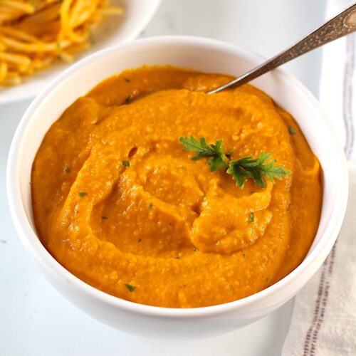 Carrot Sauce in a bowl with a spoon and parsley garnish. In background is a plate of pasta mixed with carrot sauce and a napkin on counter.