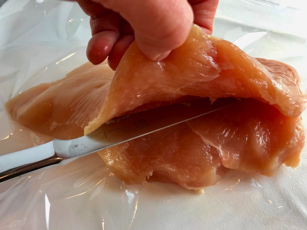 Knife starting to butterfly a chicken breast by cutting from the side while hand holds up the top half for Mozzarella stuffed chicken.