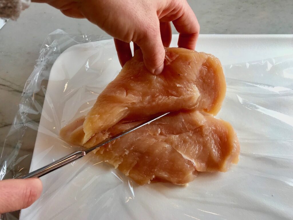 Knife cutting a chicken breast from the side while hand holds up the top half for Mozzarella stuffed chicken.