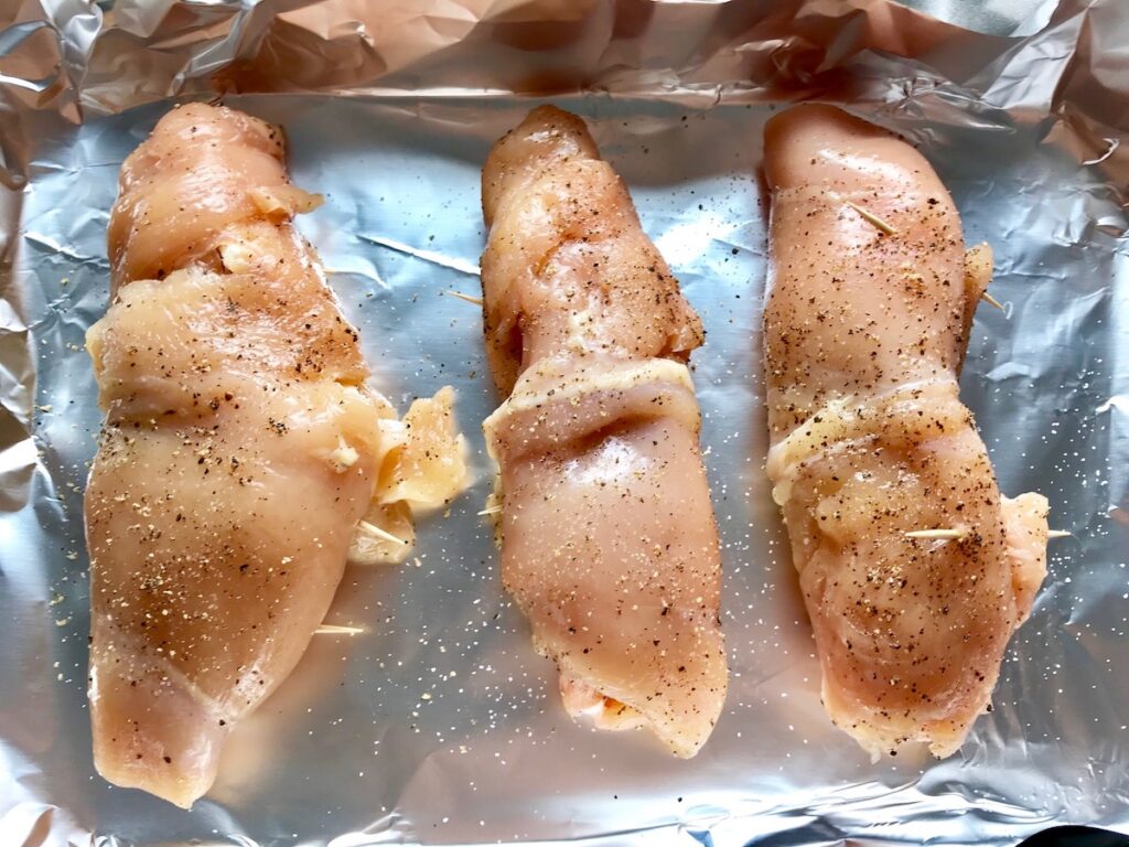 Raw rolled chicken with toothpicks and salt and pepper on aluminum foil on sheet pan for Mozzarella Stuffed chicken.