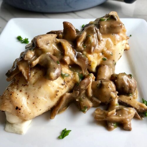 Mozzarella stuffed chicken roll on a plate with Mushroom sauce poured over top and parsley garnish sprinkled on top. In the background is the skillet with the rest of the recipe and a wood spatula.