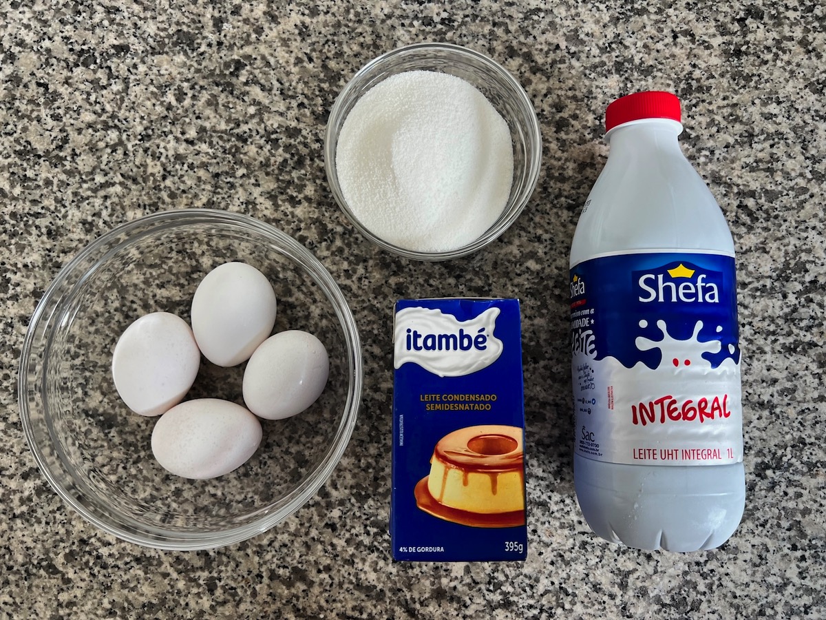 Ingredients on counter for Brazilian Flan recipe, 4 eggs in a bowl, sugar in a bowl, box of sweetened condensed milk, and milk jug.