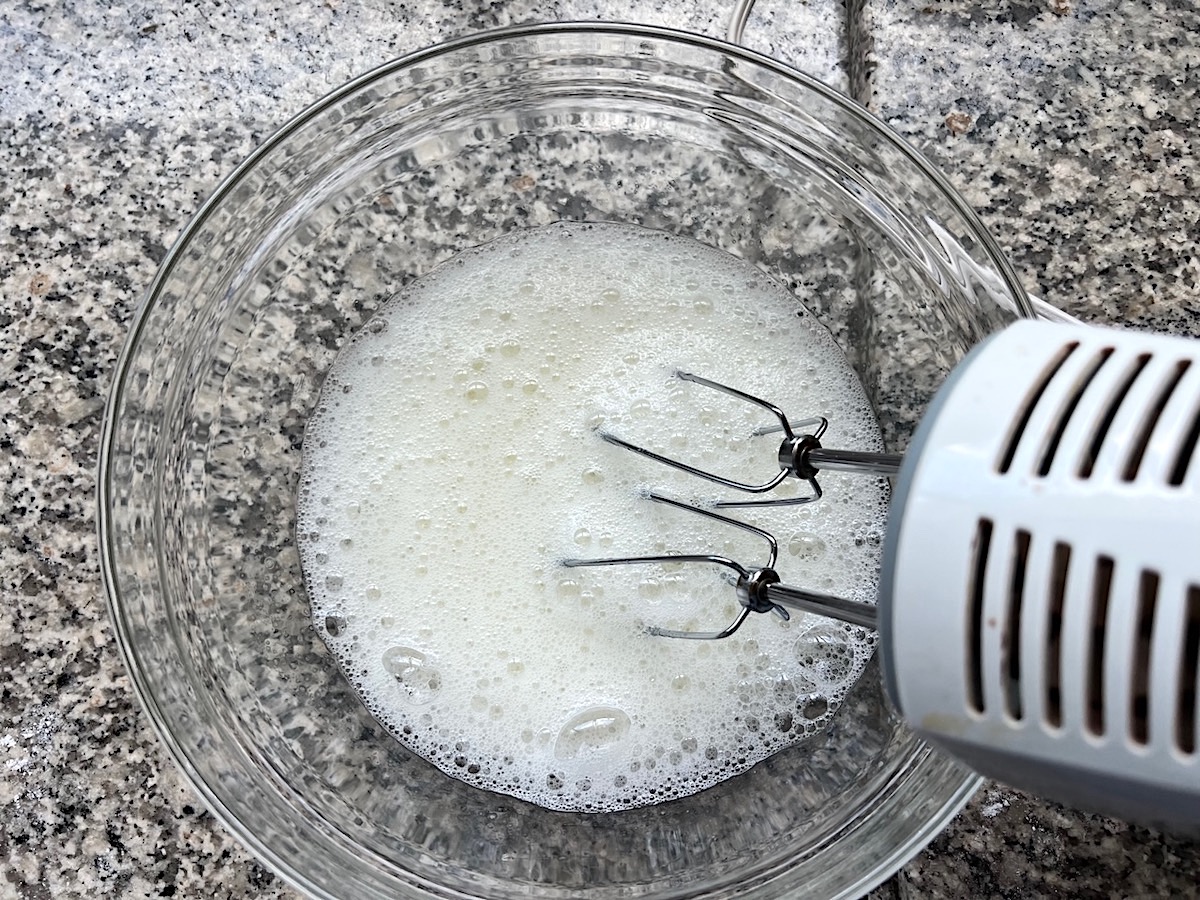 Hand mixer beating egg whites in a clear glass bowl for Brigadeiro Brazilian Cake.