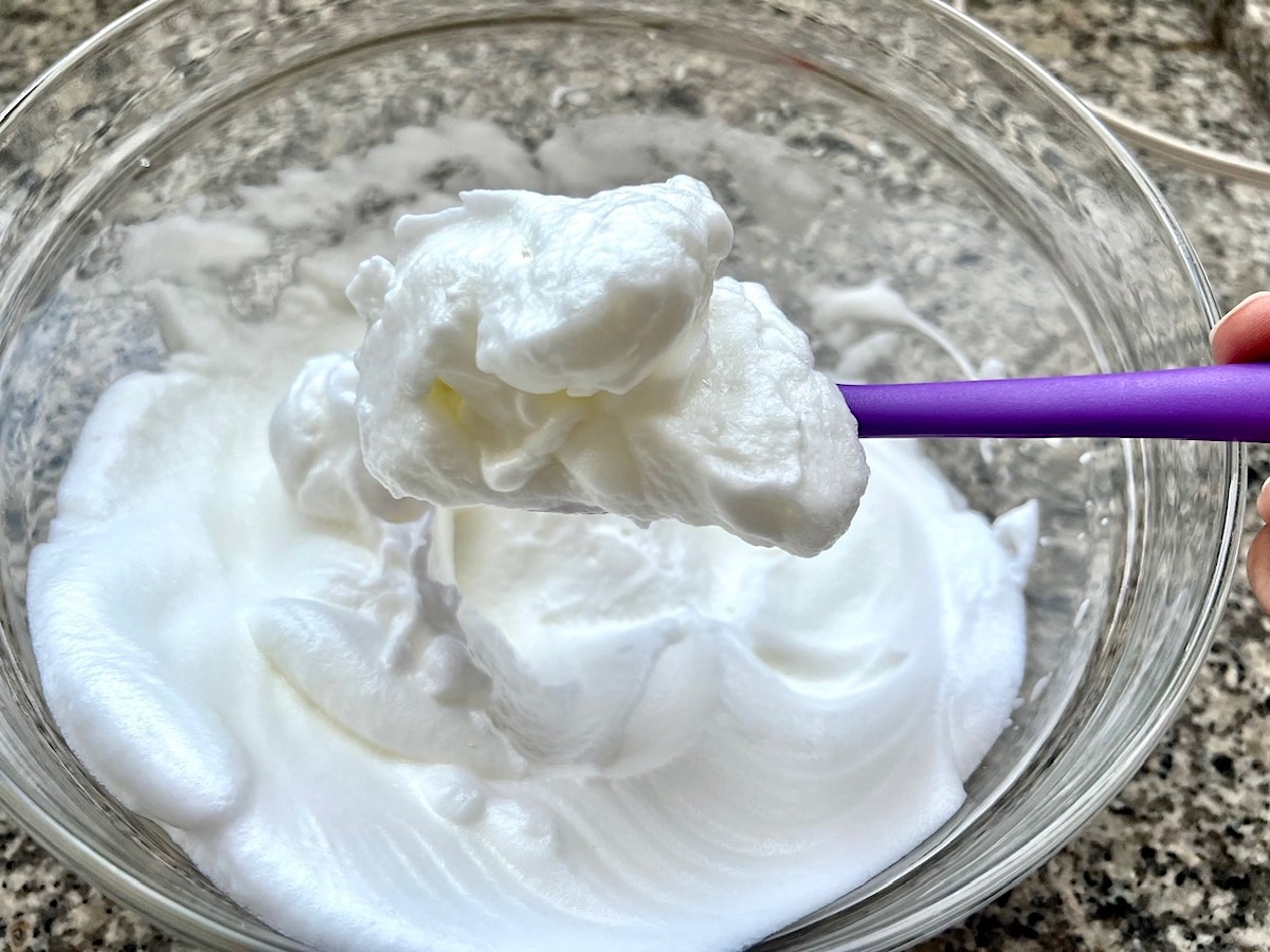 Spatula holding egg whites that have been beaten to stiff peaks in a clear glass bowl for Brigadeiro Brazilian Cake.