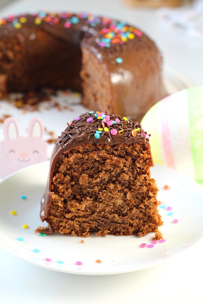 Brigadeiro Brazilian Cake slice standing up on a plate with colored sprinkles on cake and on plate. In the background is the rest of the cake on a platter along with some easter decorations.