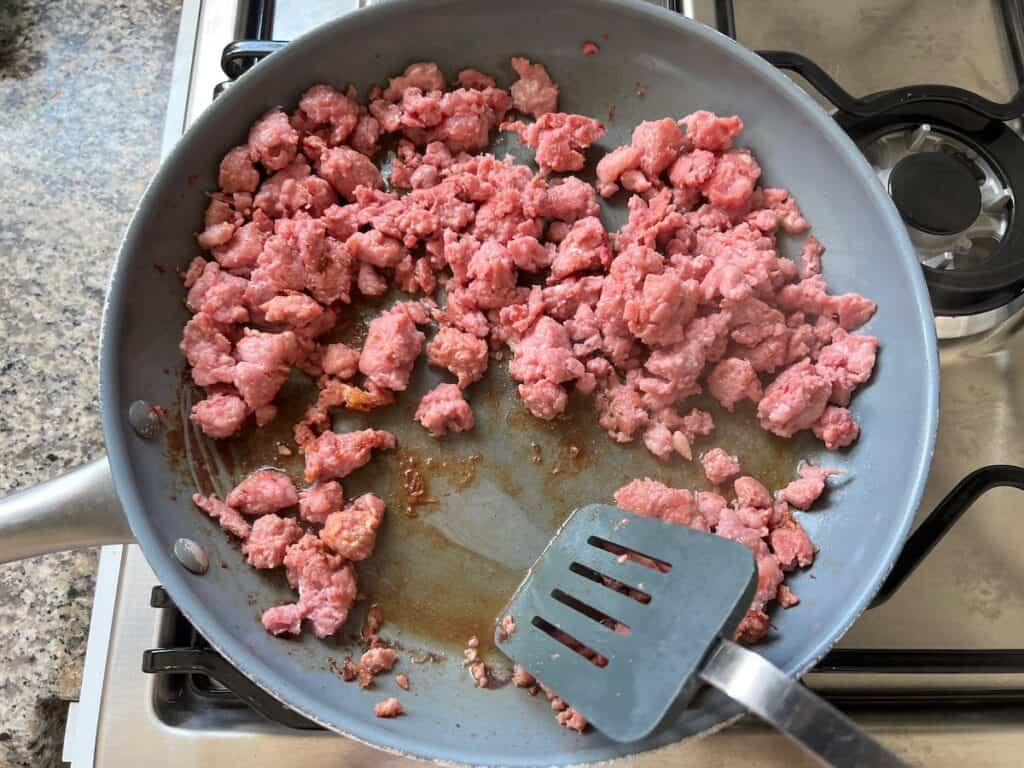 Spatula scraping up the browned bits from ground pork cooking in a frying pan for Ground Pork Slow Cooker Casserole.