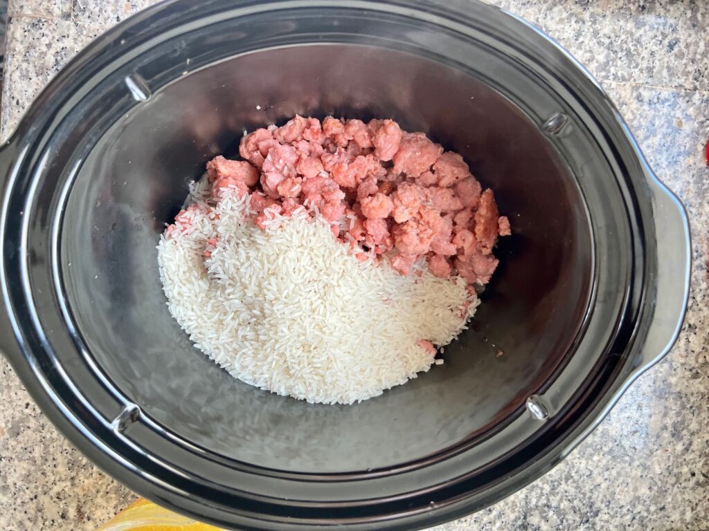 Crockpot insert on counter with raw ground port and uncooked rice added for Ground Pork Slow Cooker Casserole.