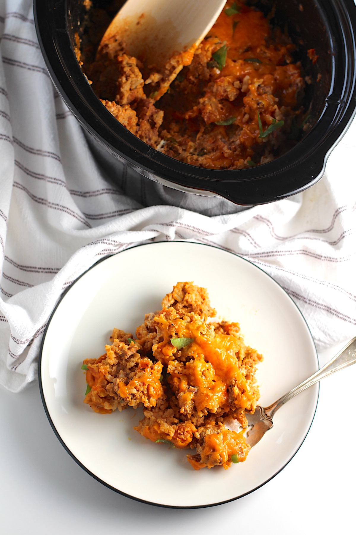Plate on counter with Ground Pork Slow Cooker Casserole on it and melted cheese on top. A fork is on plate with a bite of the casserole scooped. In the background is the slow cooker insert with wood spoon.