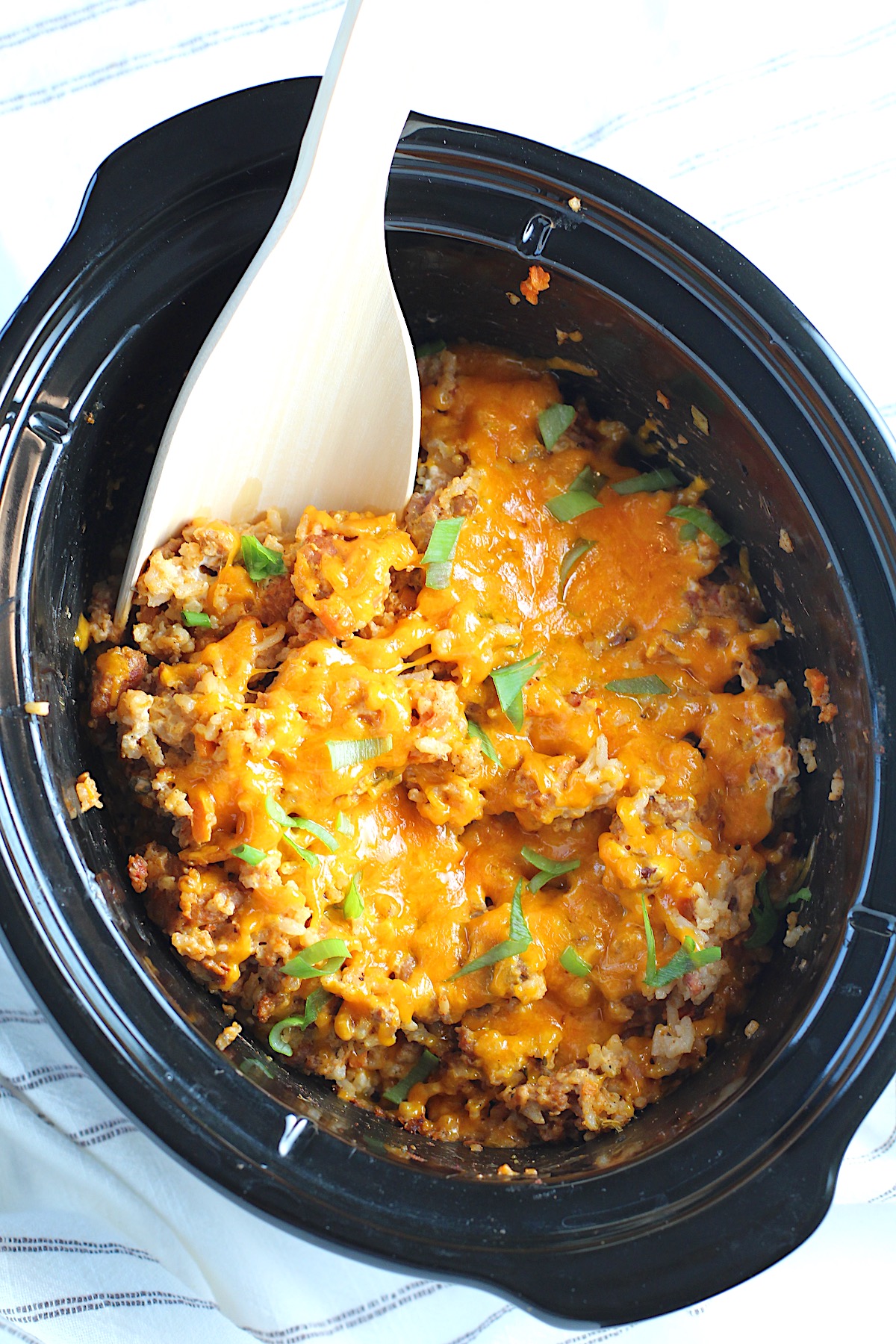 Crockpot insert on counter with Ground Pork Slow Cooker Casserole inside and melted cheese on top with scallions garnish and a wood spoon in it.