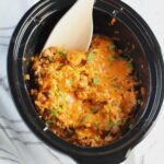 Crockpot insert on counter with Ground Pork Slow Cooker Casserole inside and melted cheese on top with scallions garnish and a wood spoon in it.
